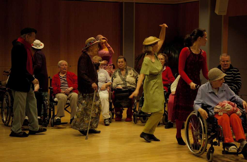 Residents of an elderly centre progressing in a dance line, other residents sitting behind them in a row