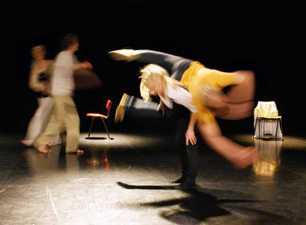 On a theatre stage, a dancer twisting around in the air on top of another dancer's back