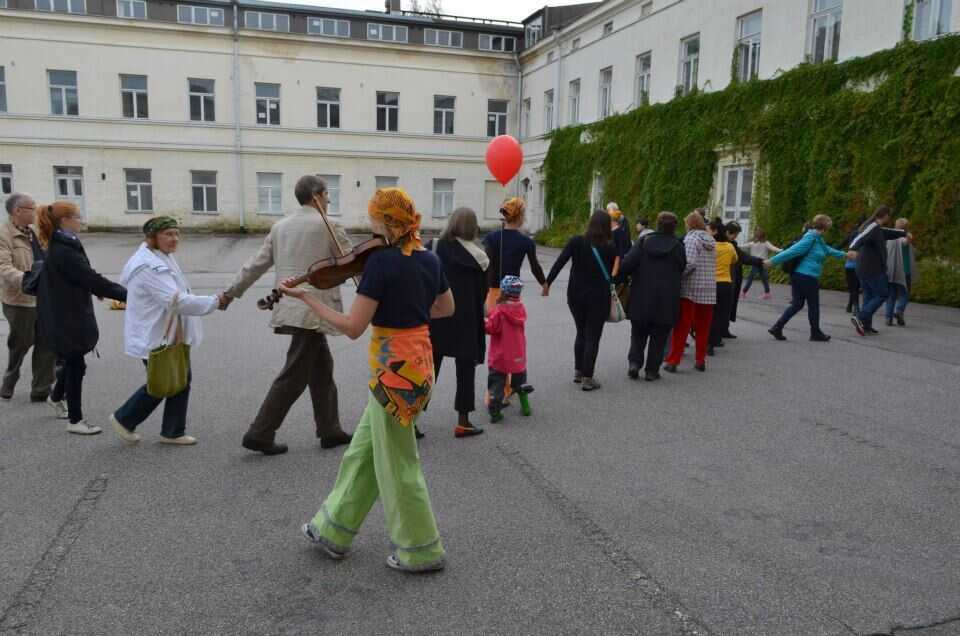 Ihmisiä kävelee käsi kädessä kiemurtelevassa letkassa ison valkoisen rakennuksen pihalla. Etualalla soittaa viulisti värikkäissä vaatteissa.