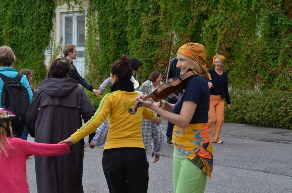 Ihmisiä kävelee käsi kädessä kiemurtelevassa letkassa muratin peittämän rakennuksen pihalla. Vieressä soittaa viulisti värikkäissä vaatteissa.