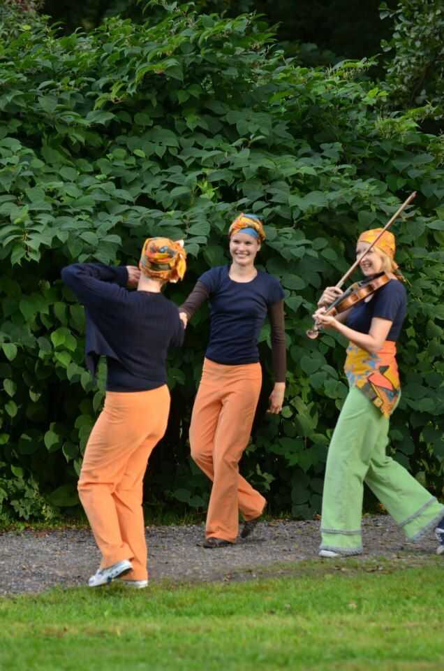 Kaksi tanssijaa ja viulisti värikkäissä vaatteissa etenevät hiekkatiellä puistossa.