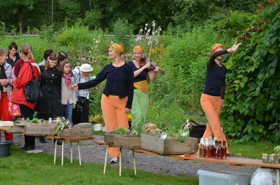 Kaksi tanssijaa ja viulisti värikkäissä vaatteissa johdattavat ihmisletkaa puistossa. Etualalla laatikoissa sadonkorjuutuotteita.