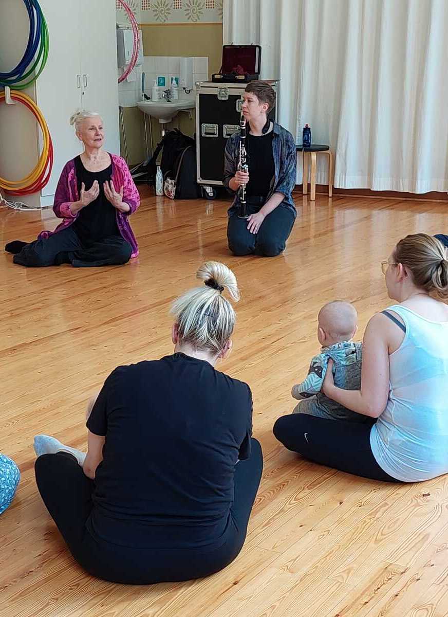 Aikuisia istumassa piirissä tanssisalin lattialla, osalla vauva sylissään. Yksi aikuisista ohjaa ja elehtii käsillään.