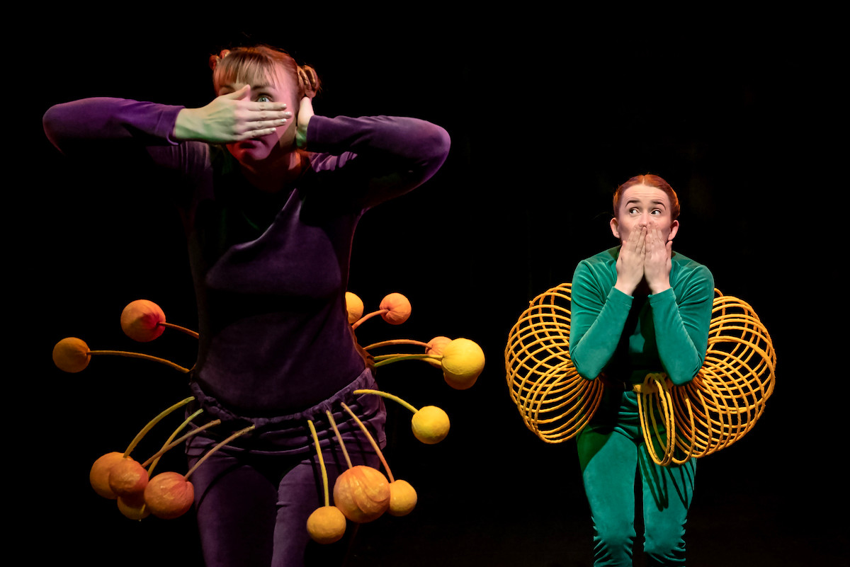 A dancer in a purple costume is covering her eyes and ears with her hands. Next to her a dancer in a green costume is covering her mouth with her hands.