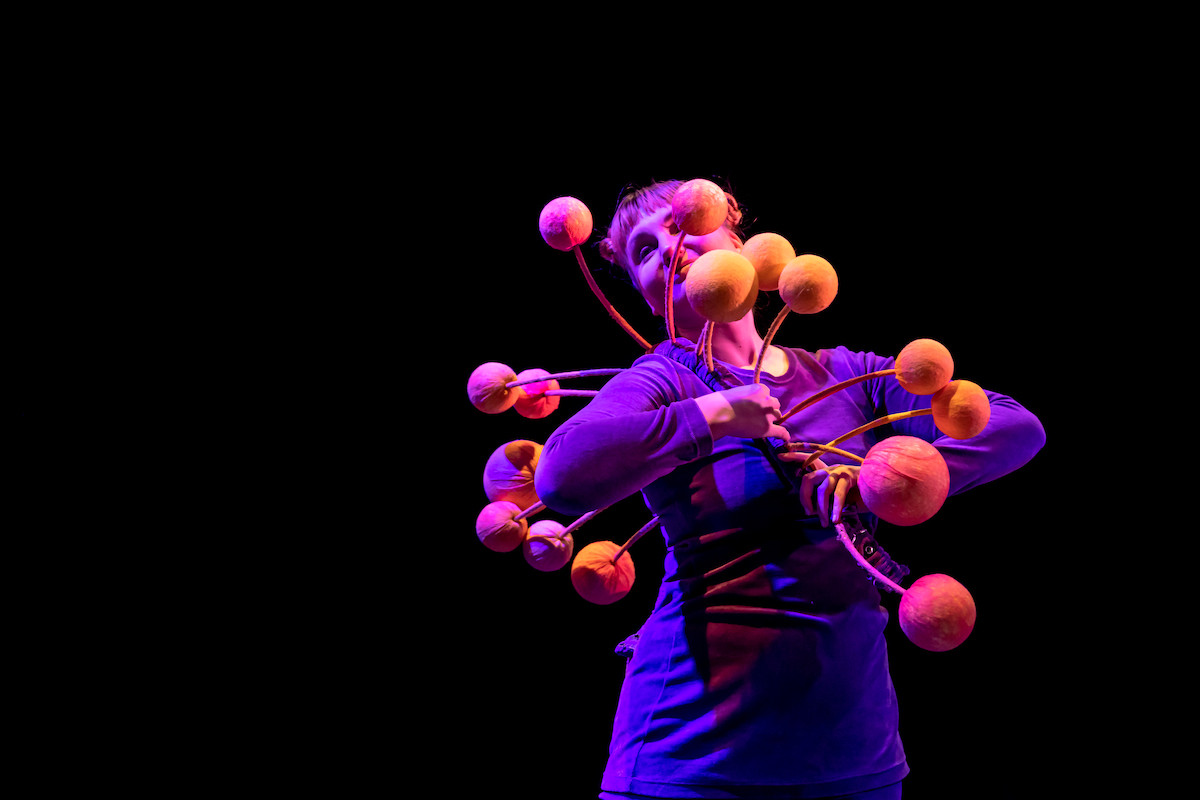 A dancer in a purple costume is dancing. She is wearing an accessory that has large orange balls attached to it via thin stems.