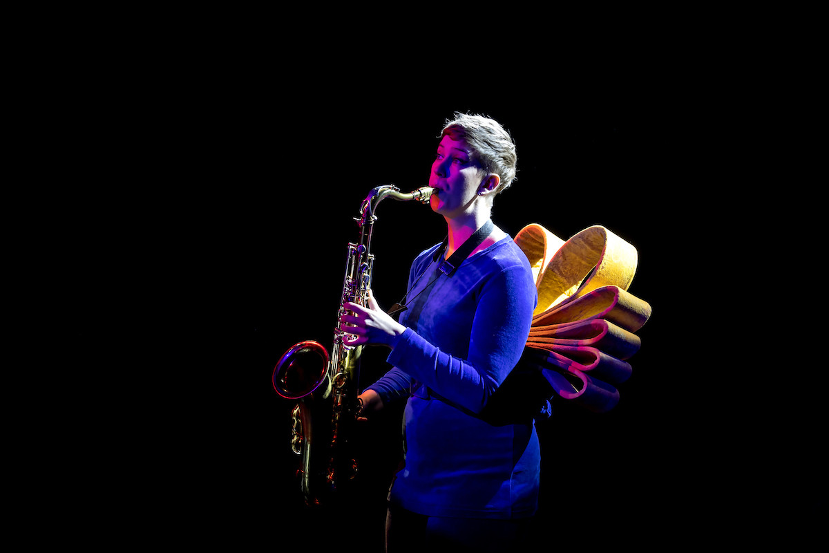 A saxophonist in a blue costume is playing. She has an accessory at the back that resembles large yellow flower petals.